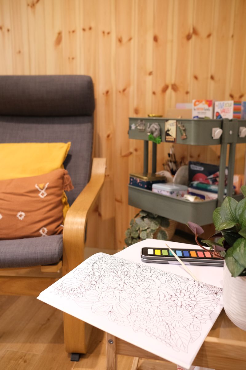 An open colouring book and watercolour paints on a small table, with an art trolley of craft supplies behind