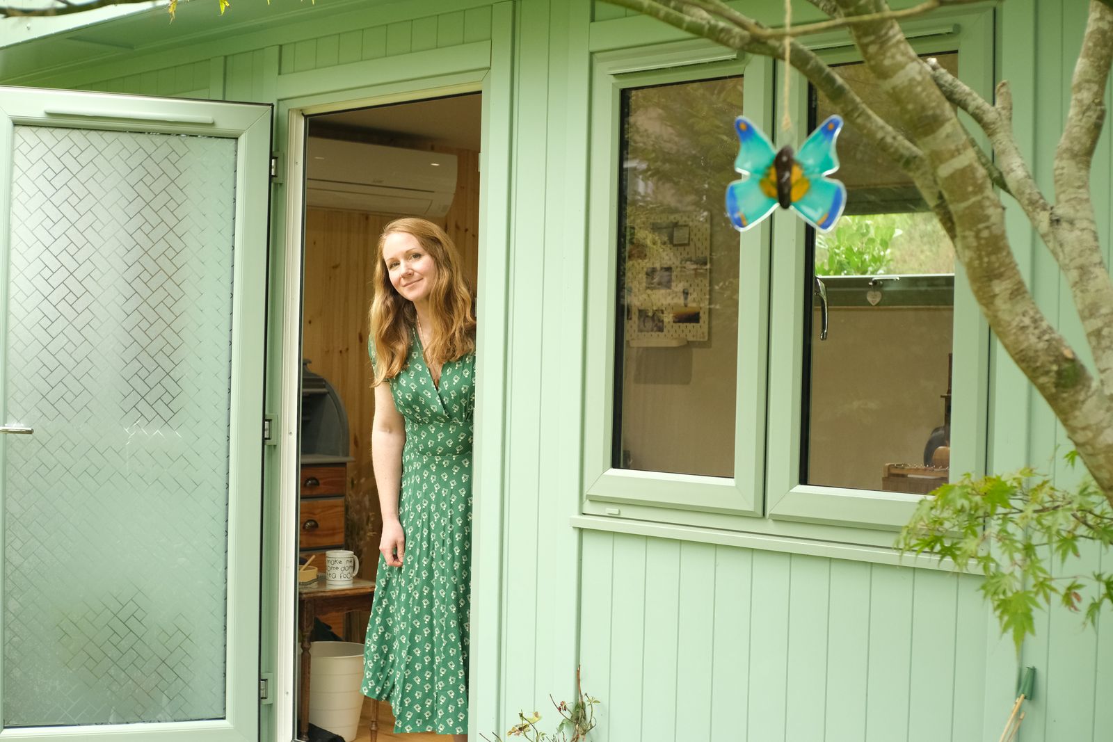 Charlotte standing in the open doorway of the sage green garden counselling room, with a glass butterfly hanging from the tree outside
